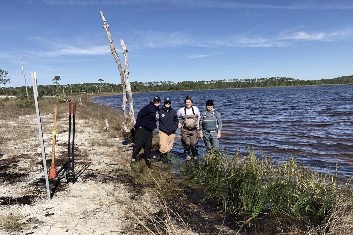 Four women stand next to newly planted vegetation along a lagoon shoreline.